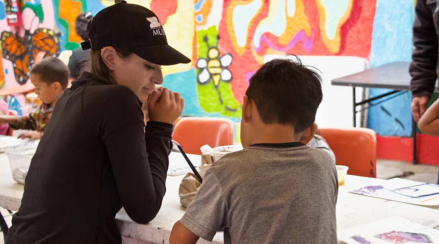 In a room with colorful art painted on the walls, a woman sits to the right of a young boy as he paints.